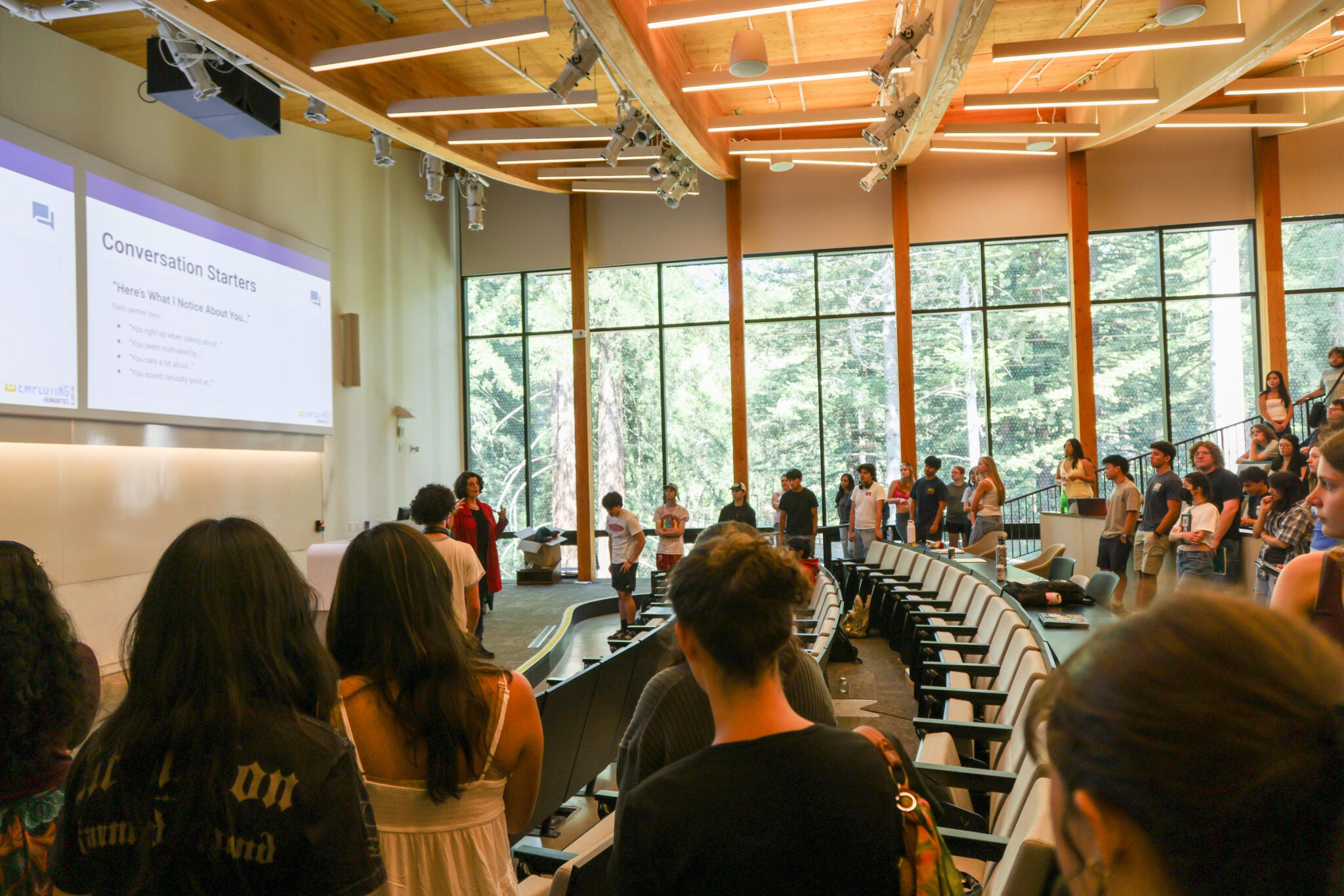 Students stand in a large lecture hall with redwoods visible through the windows