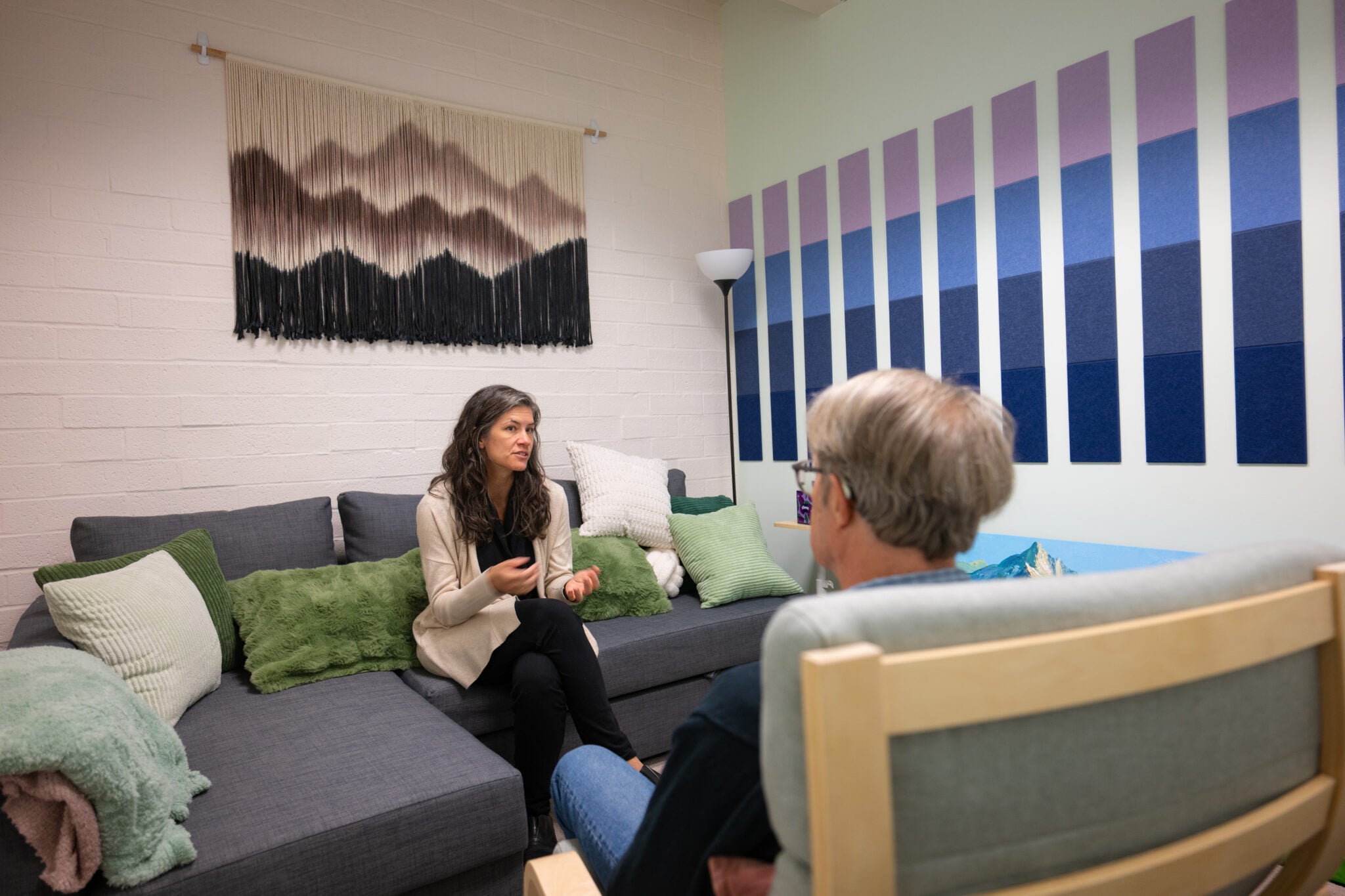 Two people sit across from one another in a brightly lit room that is furnished with a comfortable couch and chair and colorful art on the wall