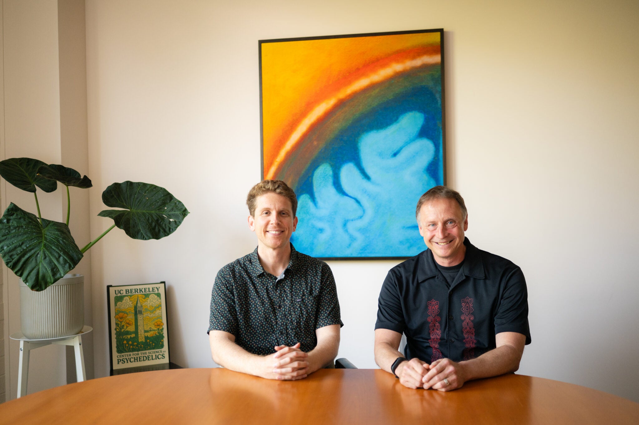 Two people sit at a table smiling at the camera. Behind them is a painting with blue swirls that are reminiscent of brain matter