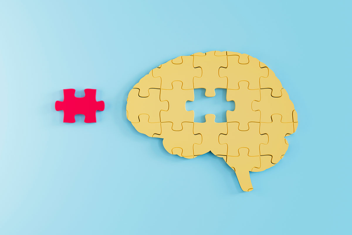 A yellow jigsaw puzzle in the shape of a brain, with one missing piece. The missing piece is red and sits beside the puzzle.