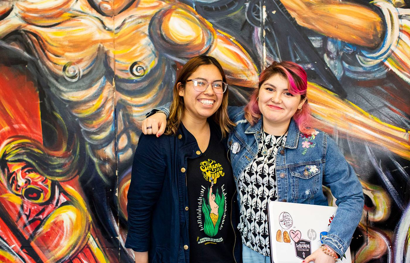 Two students smiling in front of a large mural with gold, black and red coloring.
