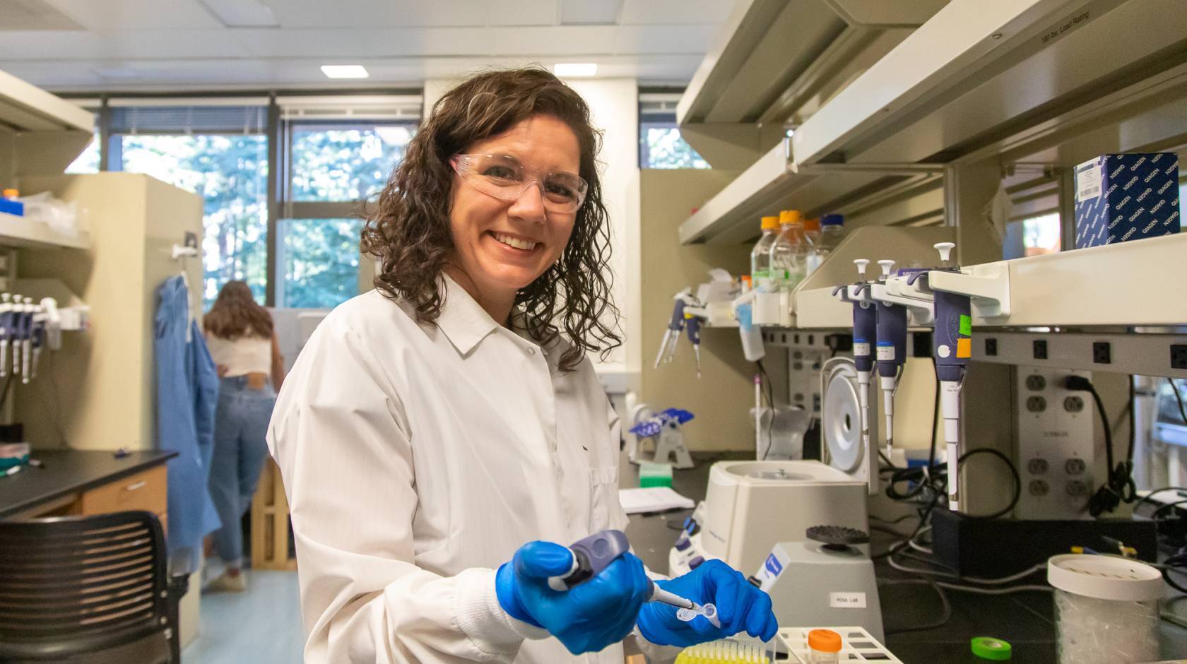 Karen Miga in safety glasses, white coat and blue gloves looks up from pipetting to smile at the camera in a lab