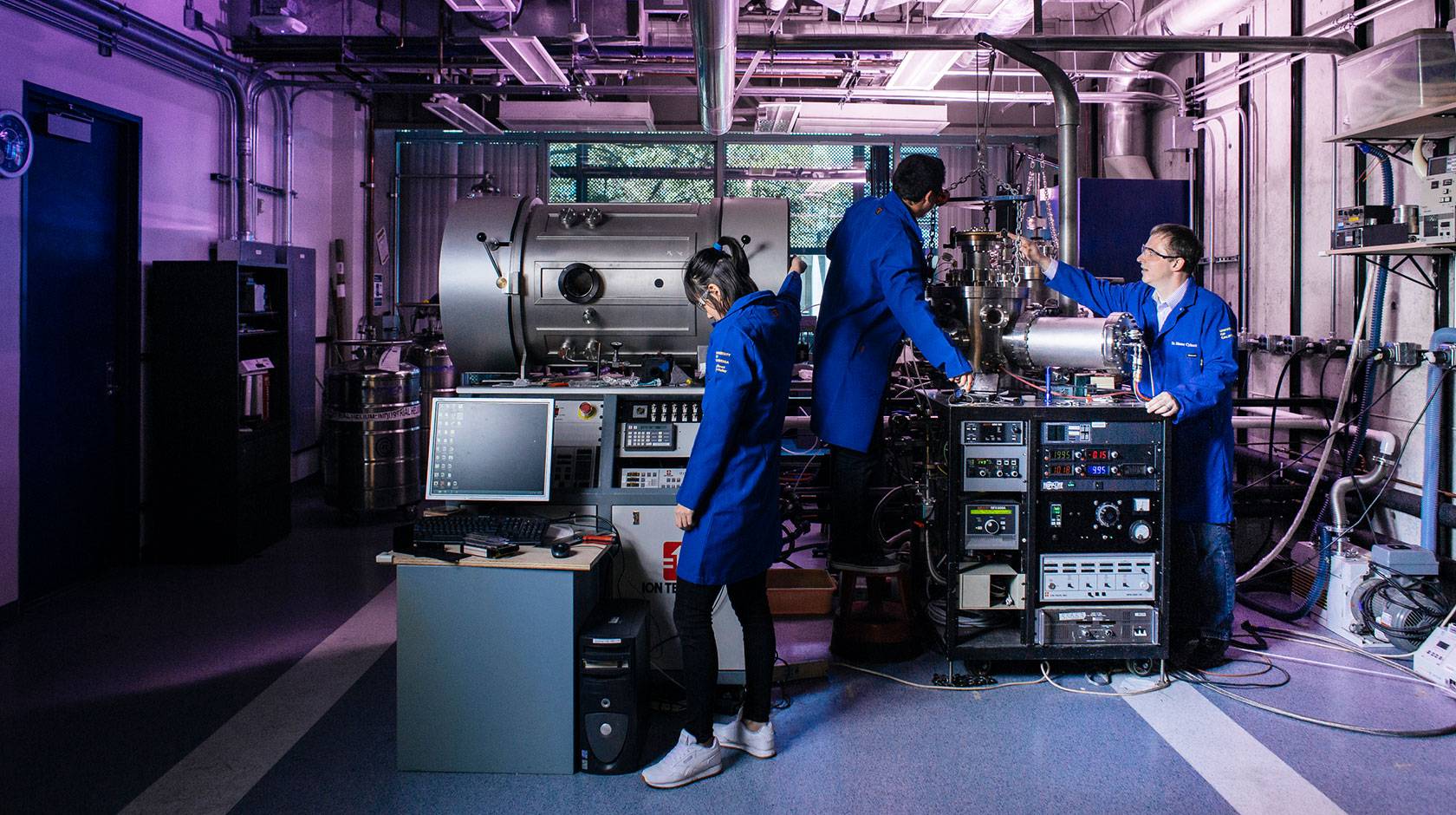 Three people in blue lab coats work in a purple-lit facility with large, complex-looking equipment.