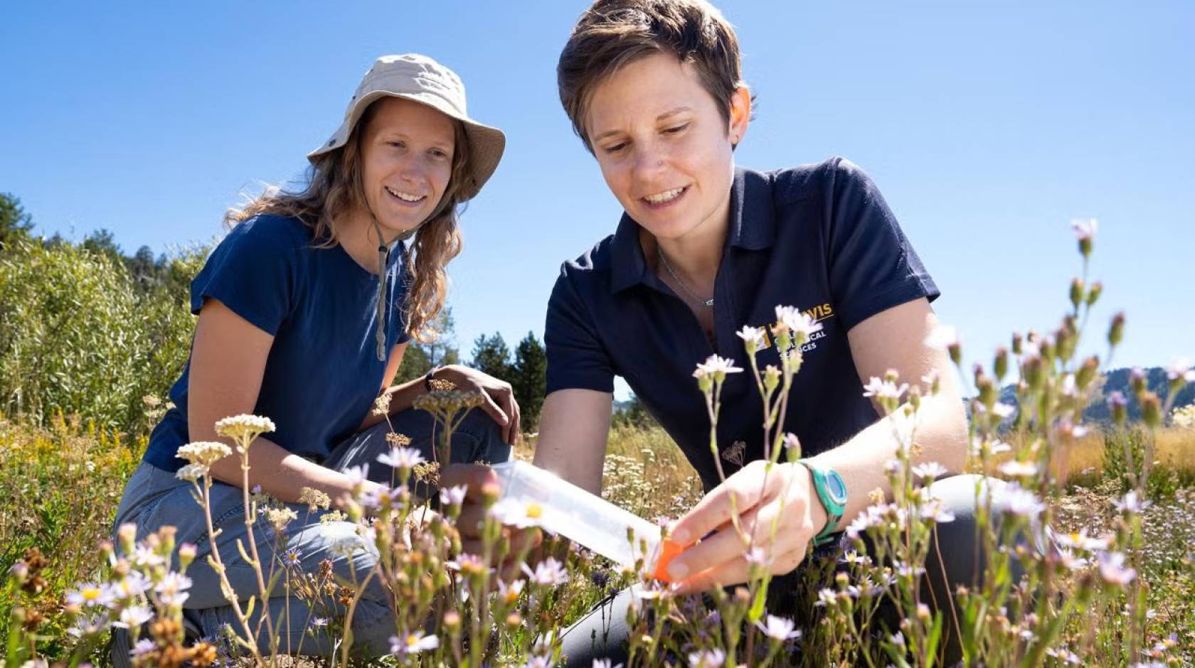 Two women in dark blue shirts kneeling in a meadow. One is holding a plastic tube while the other looks on