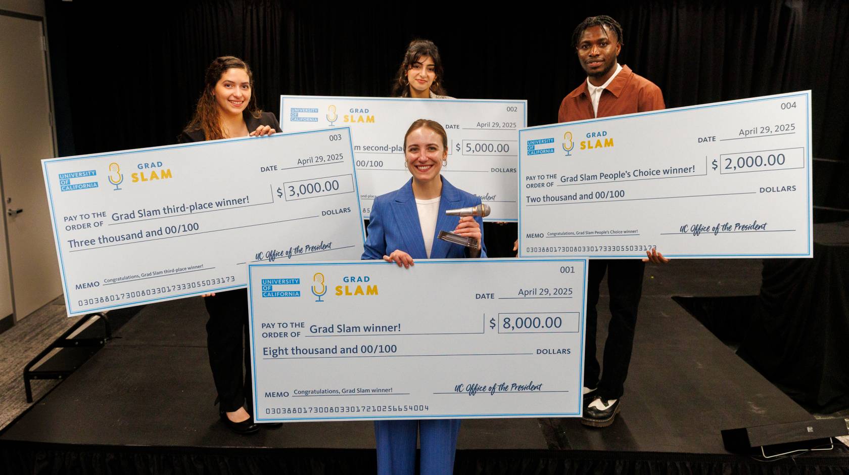 Four people, one in the foreground, three in the background, hold up giant checks