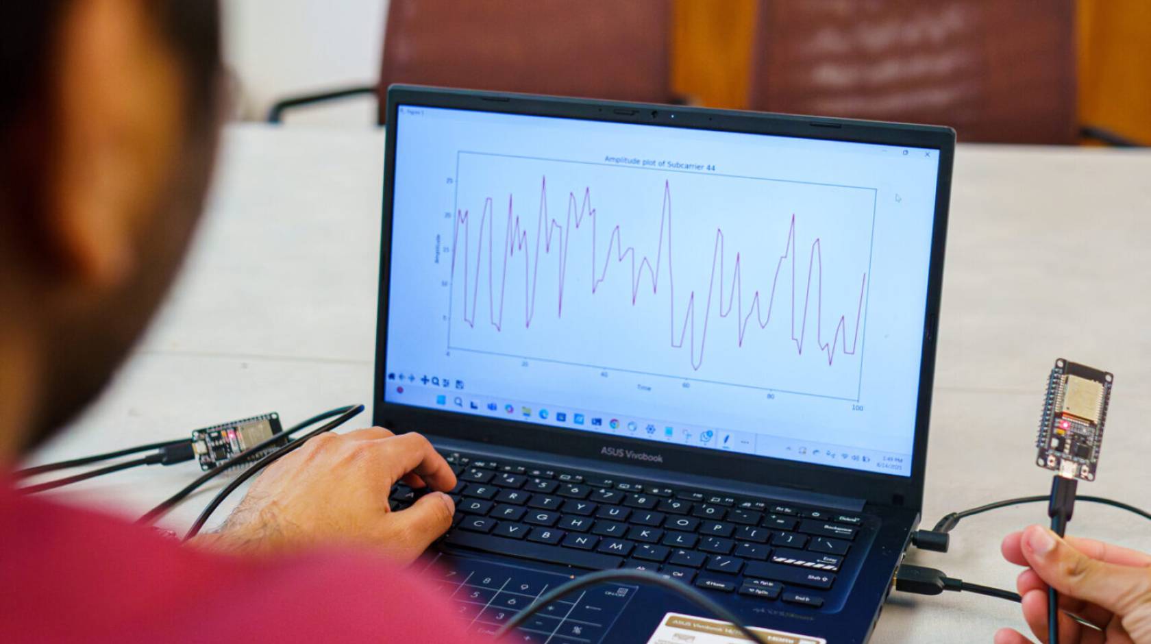 A researcher holds a wifi device in front of a laptop that displays heart rate information.