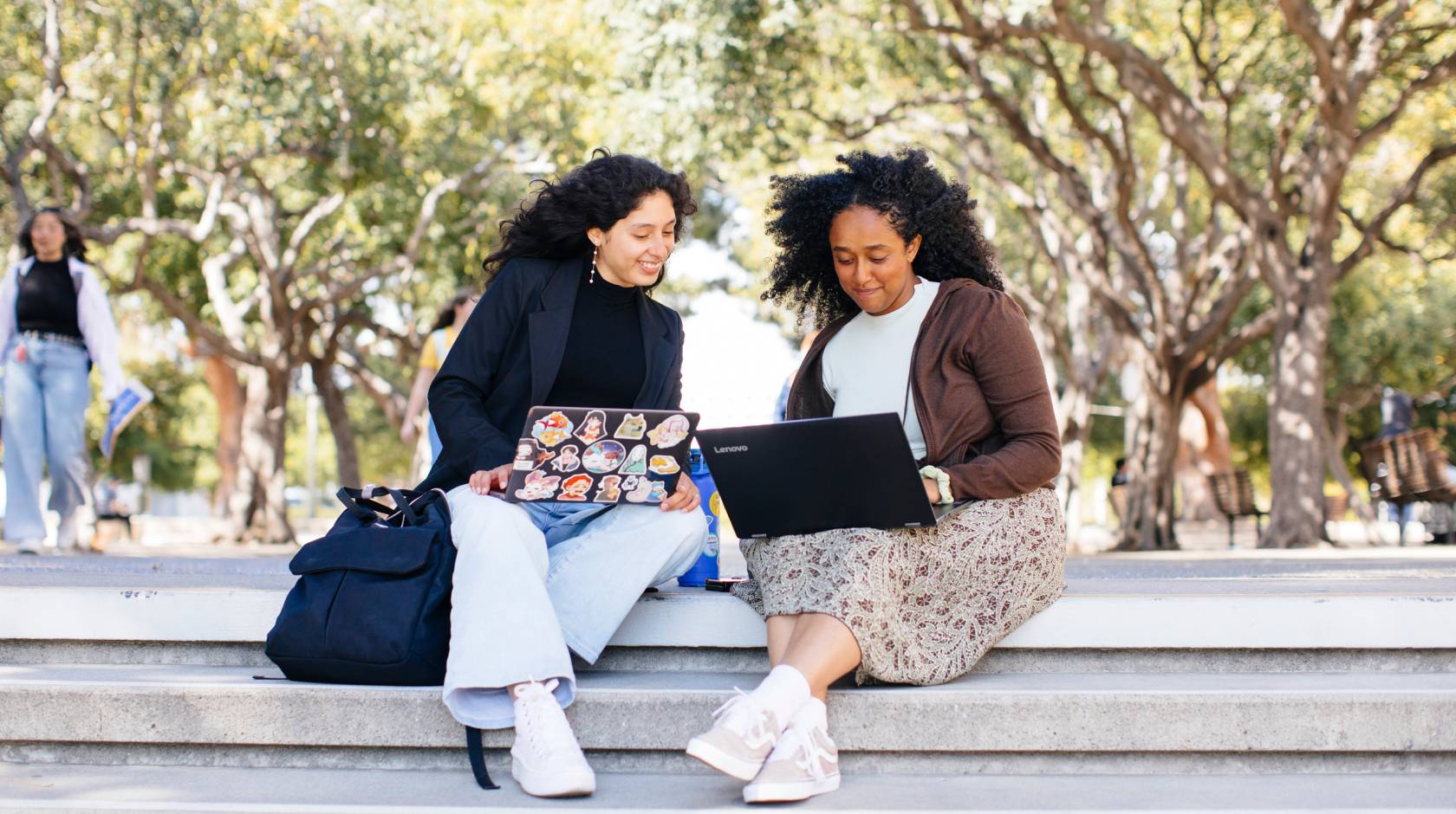 Two students sitting on a stairway outside looking at laptops together and smiling