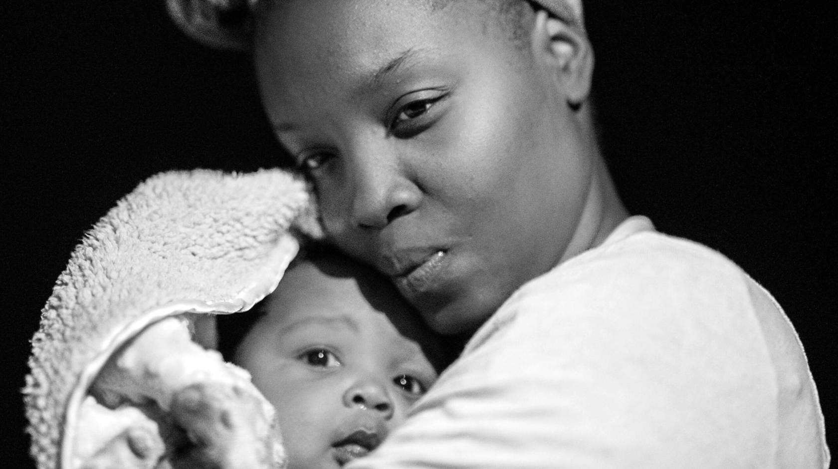 In a black-and-white close-up photo, a Black woman gazes thoughtfully as she coddles a baby wrapped in a blanket