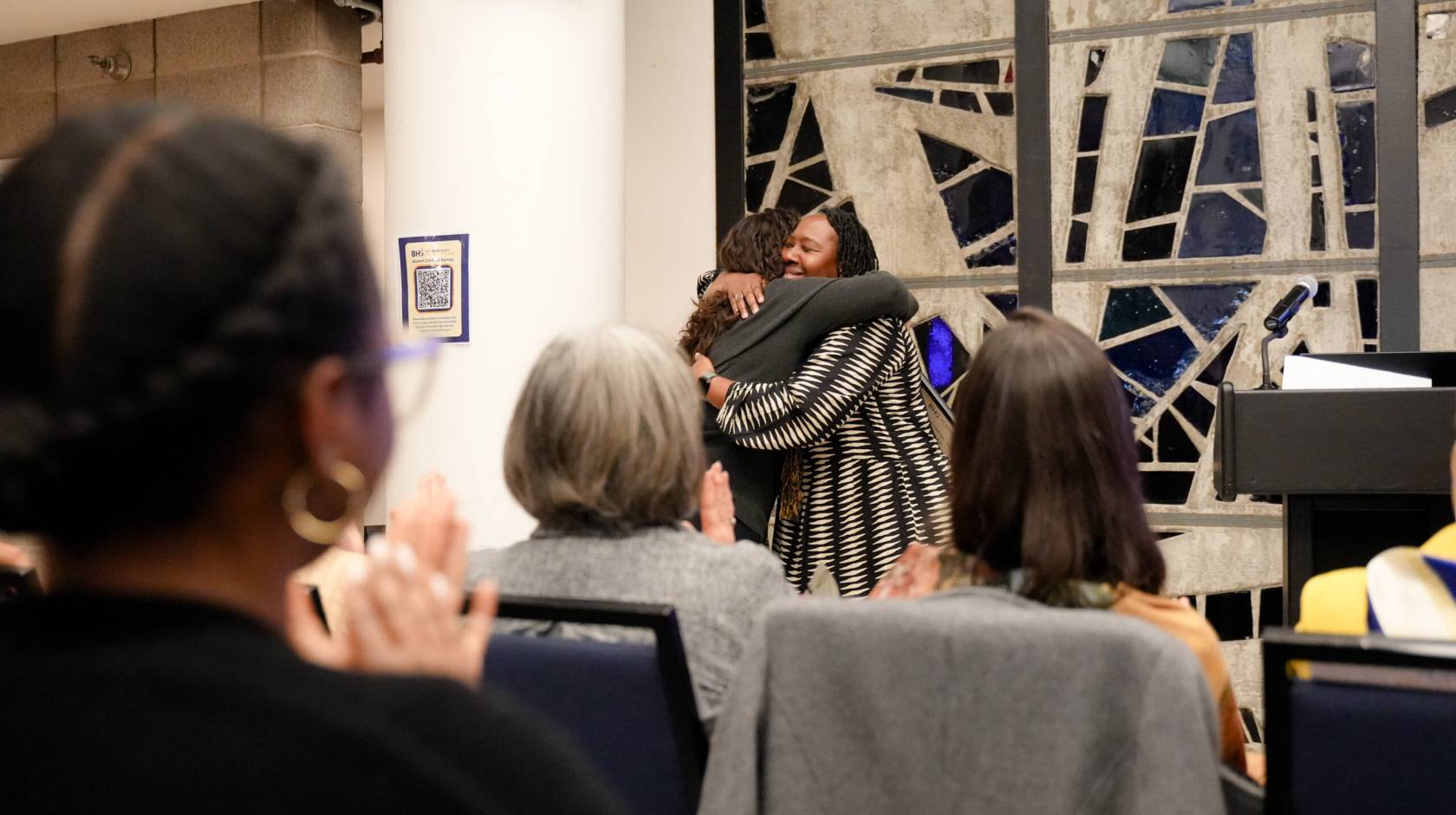 two women hug in front of a stained glass window as others applaud