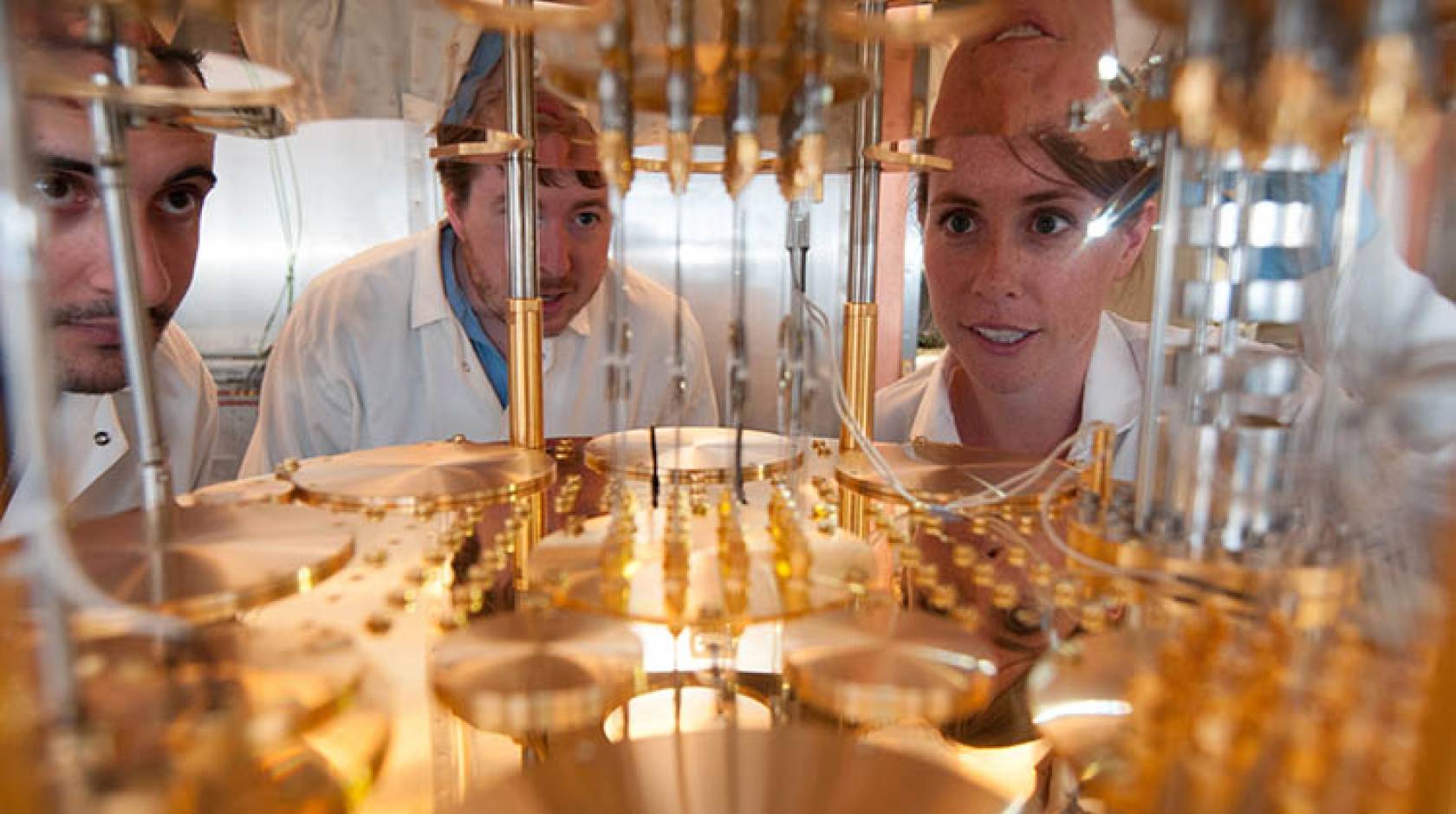 Three people in white lab coats peer into a very large, delicate, gold-colored machine.