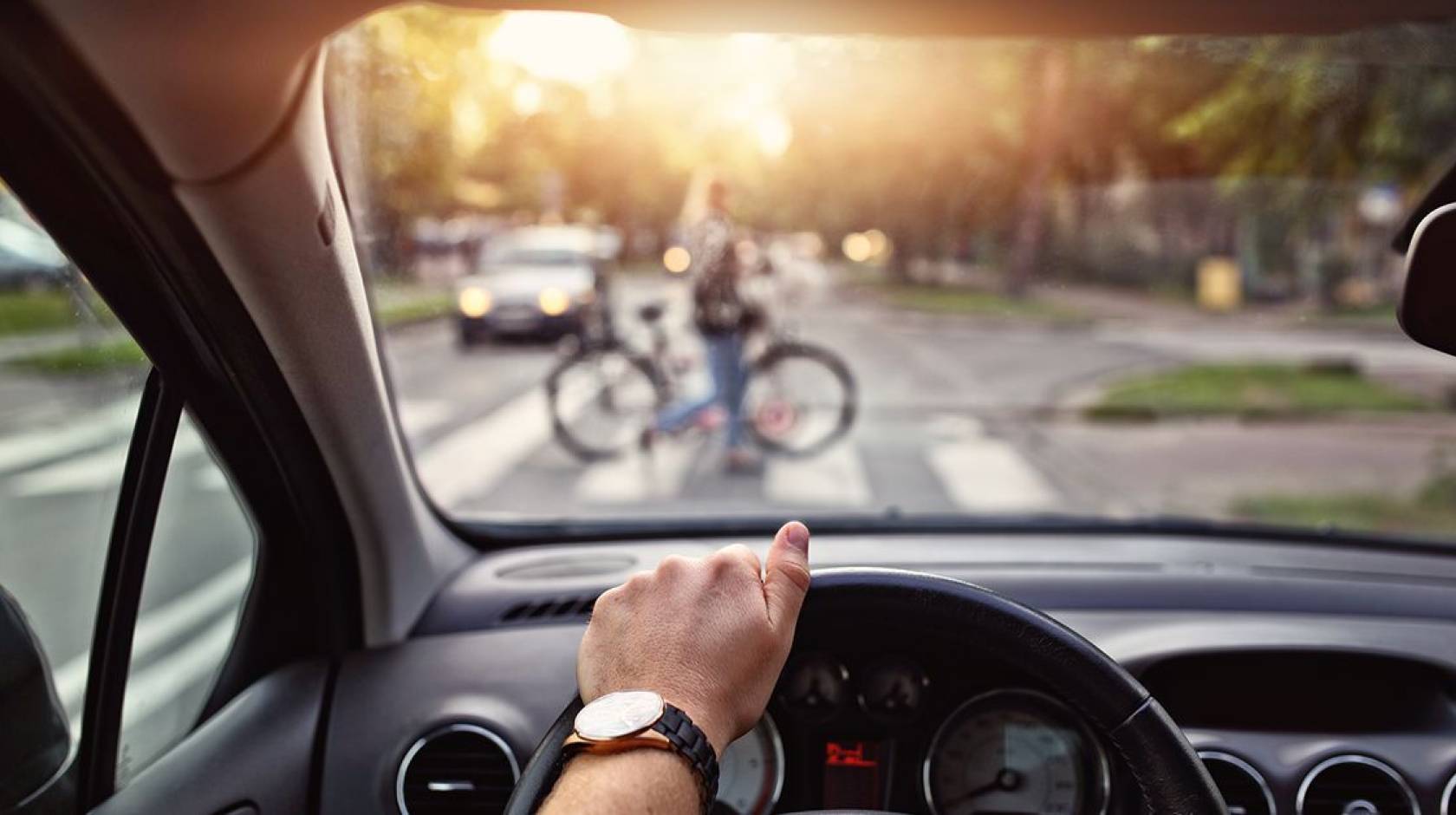 A view of someone's hand on a steering wheel and a person walking a bicycle across the crosswalk out of focus
