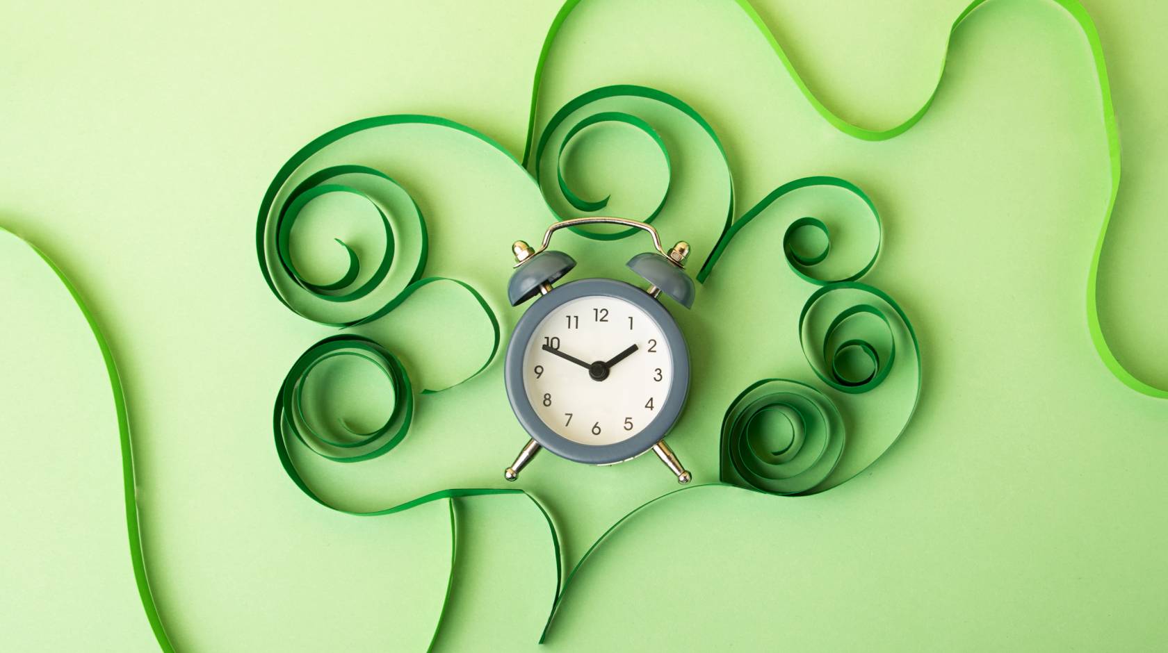 An old-fashioned alarm clock on a green background surrounded by green paper curlicues resembling a plant