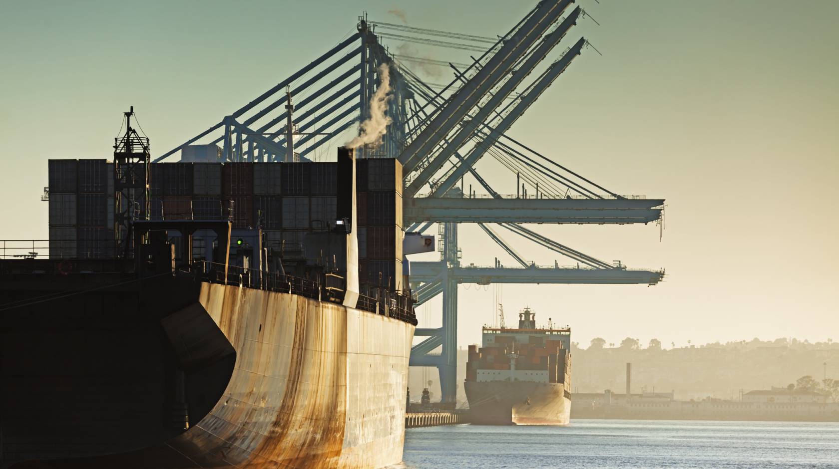 Two rusty container ships in a row, one behind, being loaded at a port during the evening