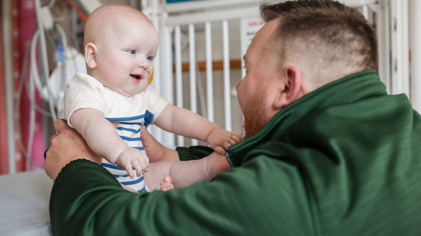 A man in a green jacket looks at a baby in front of him, both looking excitedly at each other