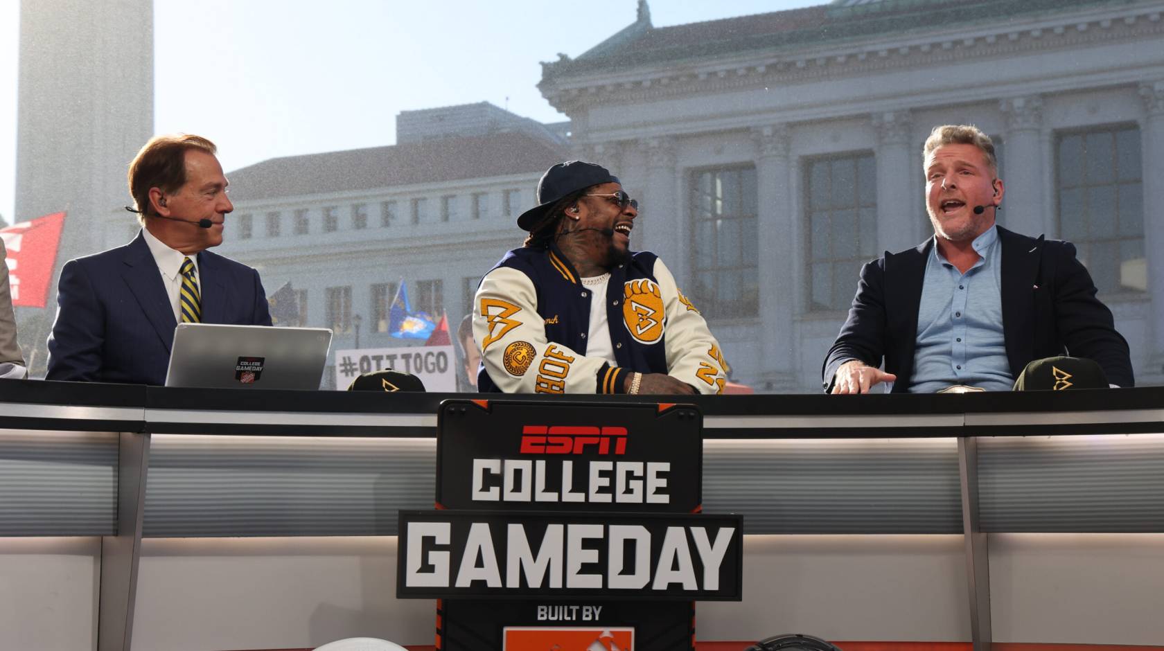 Nick Saban, man in a blue and gold tiem next to Marshawn Lynch, wearing sunglasses, a backwards hat, and a Cal bomber jacket, next to Pat McAfee, man in dress shirt and blazer without tie, mouth open, on the set of College GameDay, on the Cal campus, Campanile and Doe Library visible behind in the morning light