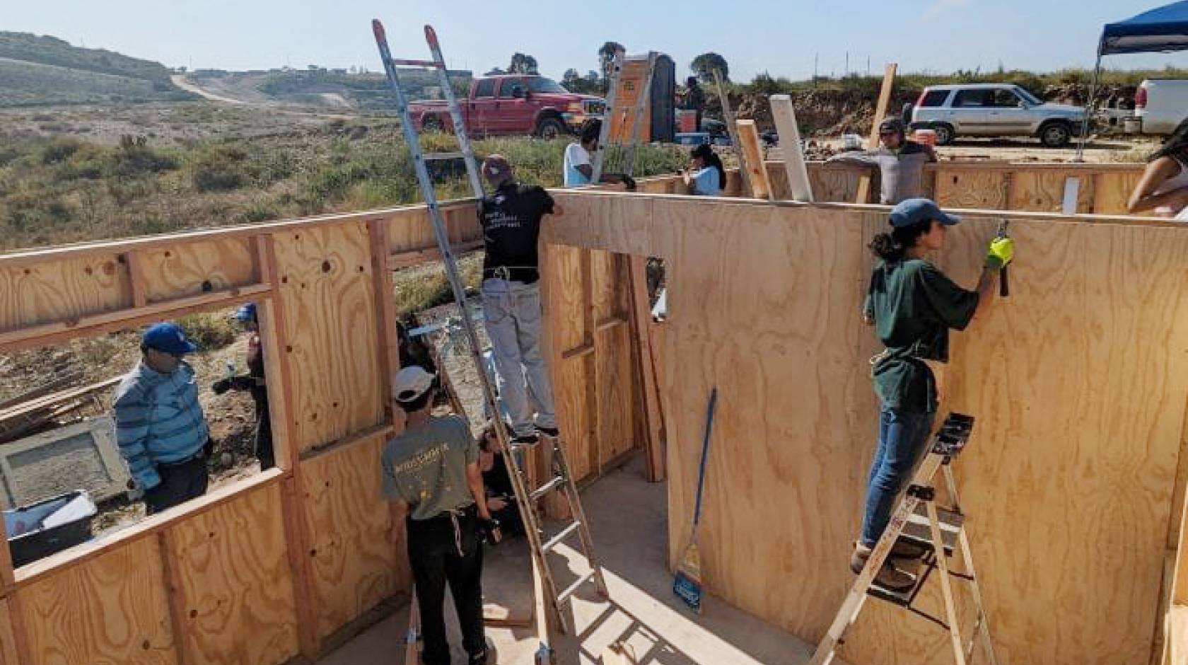 workers, some on ladders, building a house in an unpopulated, scrubby landscape