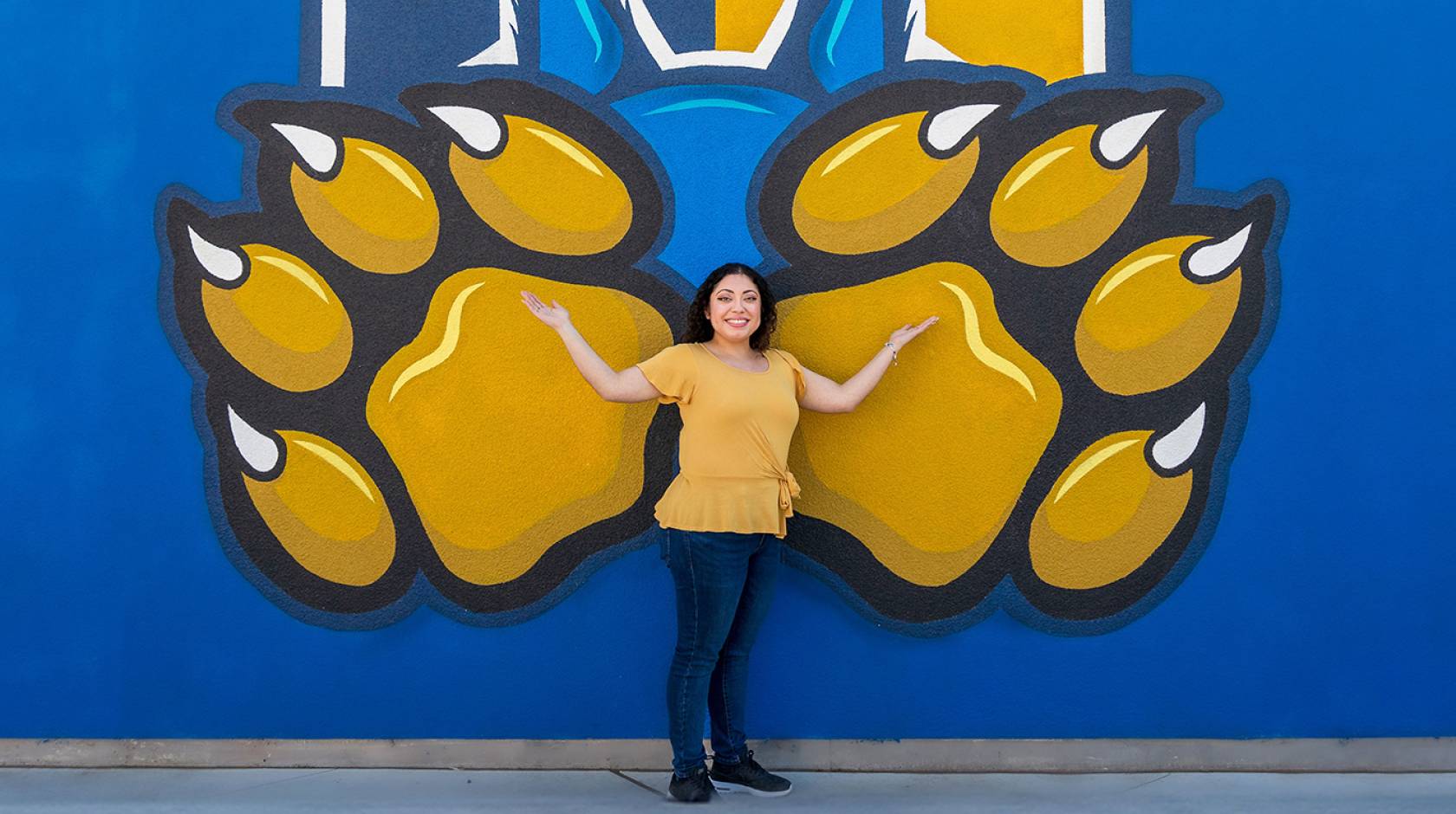 A smiling woman standing in front of a blue wall painted with the UC Merced gold bobcat paws