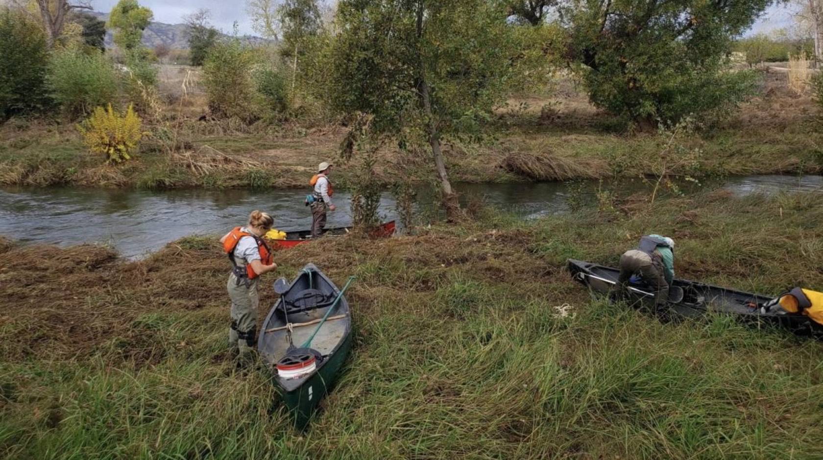Three people with water gear and canoes on the bank of a creek