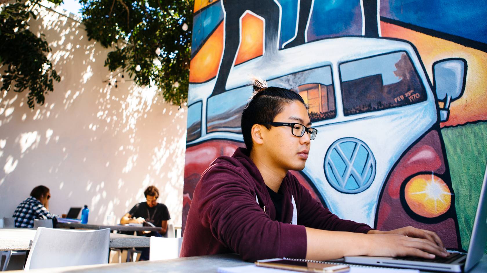 A student working on a laptop at a table outside, in front of a mural showing a Volkswagen van