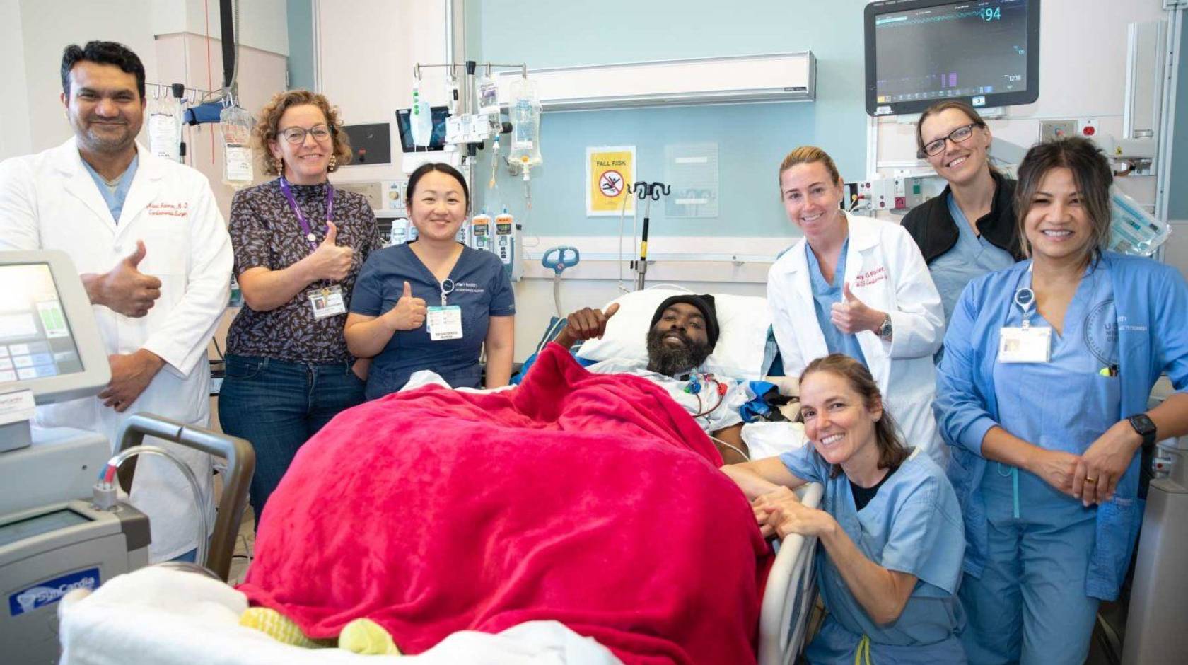 A man in a hospital bed with a red blanket on him is surrounded by a team of nurses and doctors, smiling and giving thumbs up