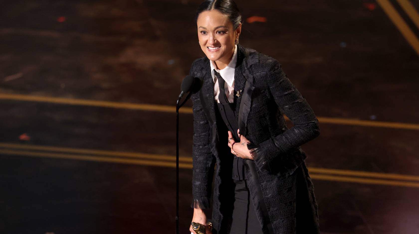 A woman speaks at a mic at the Oscars, holding the statuette and looking extremely happy