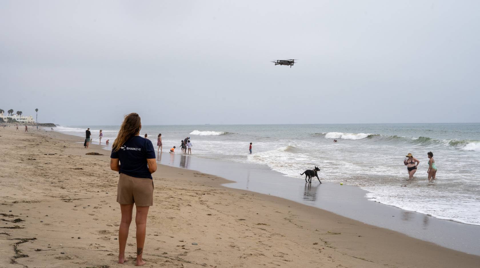 a woman stands on a busy beach looking out a a drone hovering above the waves.