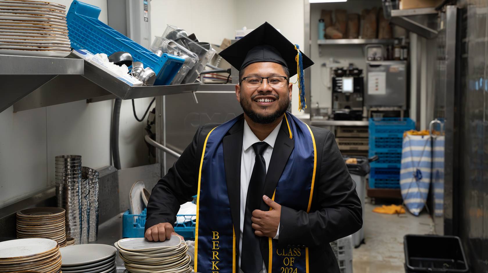 A smiling man in a college graduation cap and gown with a UC Berkeley sash stands in a commercial kitchen in front of a sink and a stack of dishes
