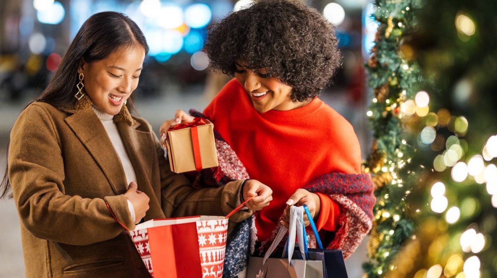 Two women smile and juggled shopping bags next to a Christmas tree.