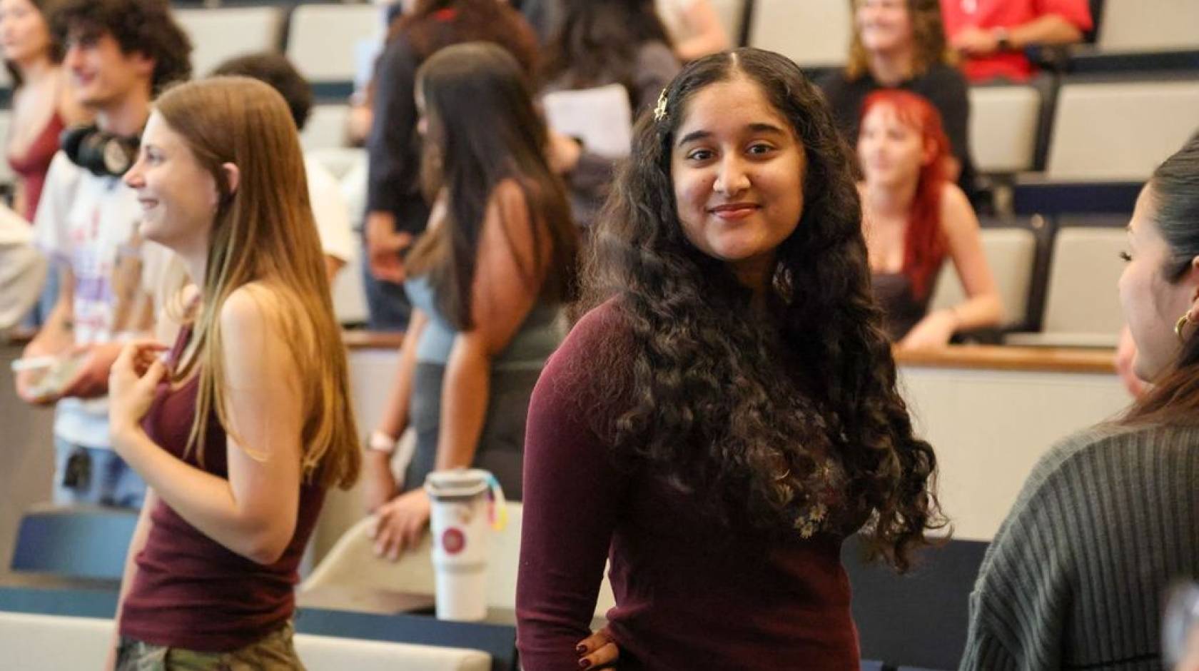 A young woman smiles at the camera with other students standing smiling looking away from the camera behind her