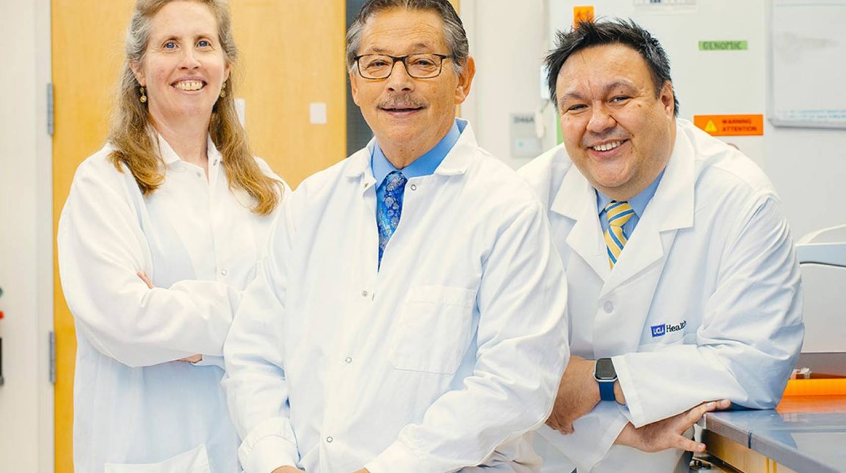 Three doctors with white lab coats pose together in a lab and smile