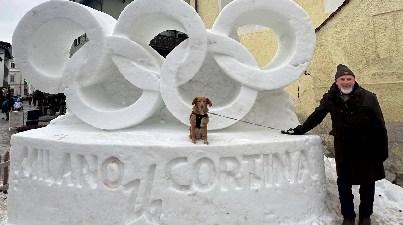 In front of a snow sculpture of the Olympic rings that reads Milano Cortina, a man stands next to a service dog sitting on the sculpture