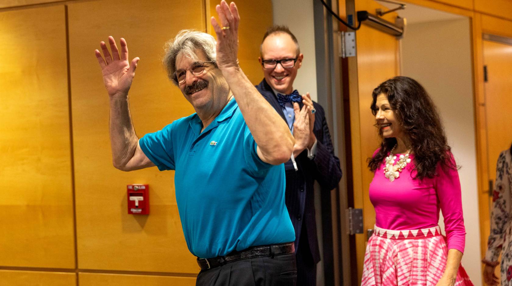 An older man with thick hair and a mustache and glasses throws up his hands as he enters a room and smiles as a woman in a pink dress follows behind, also smilling, and a man behind them smiles, looking on