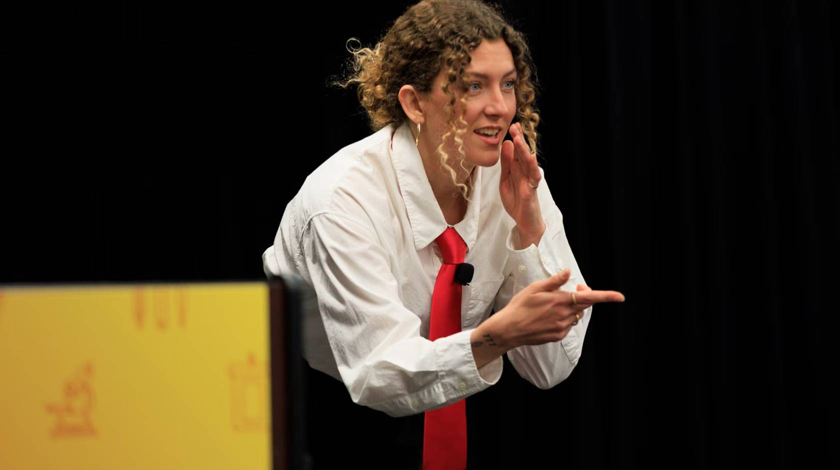 A woman in a white blouse and red tie makes a chopping gesture while pointing during a presentation