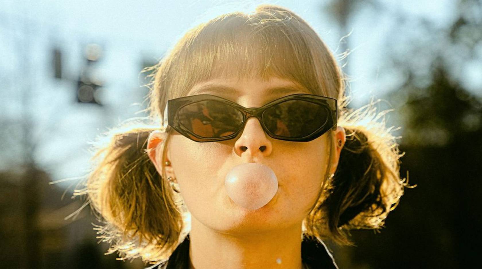 A woman wearing sunglasses and pigtails blows a gum bubble facing the camera