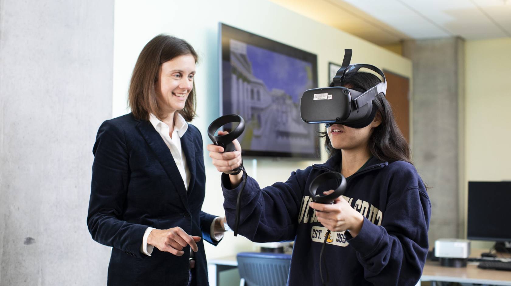 A woman in a jacket and blouse smiles next to someone in a VR headset with a sweatshirt