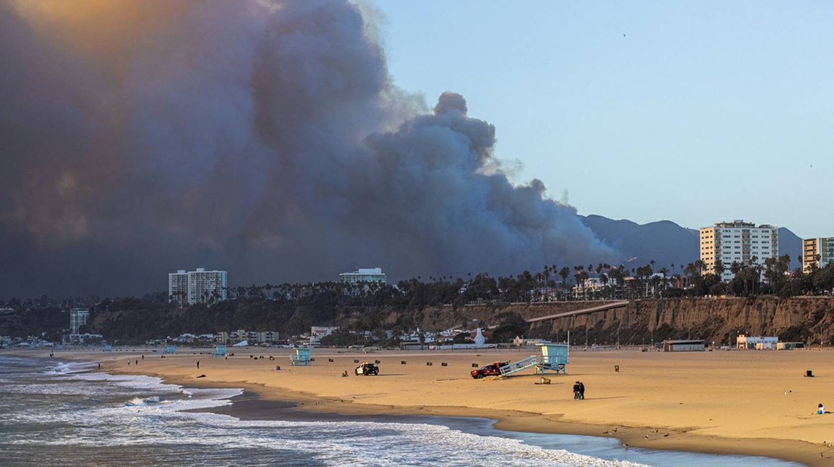 A wide sandy beach with gentle waves in the foreground, lifeguard towers and a few people scattered along the shore, while a massive dark plume of smoke rises from hills behind nearby coastal buildings under a clear blue sky.