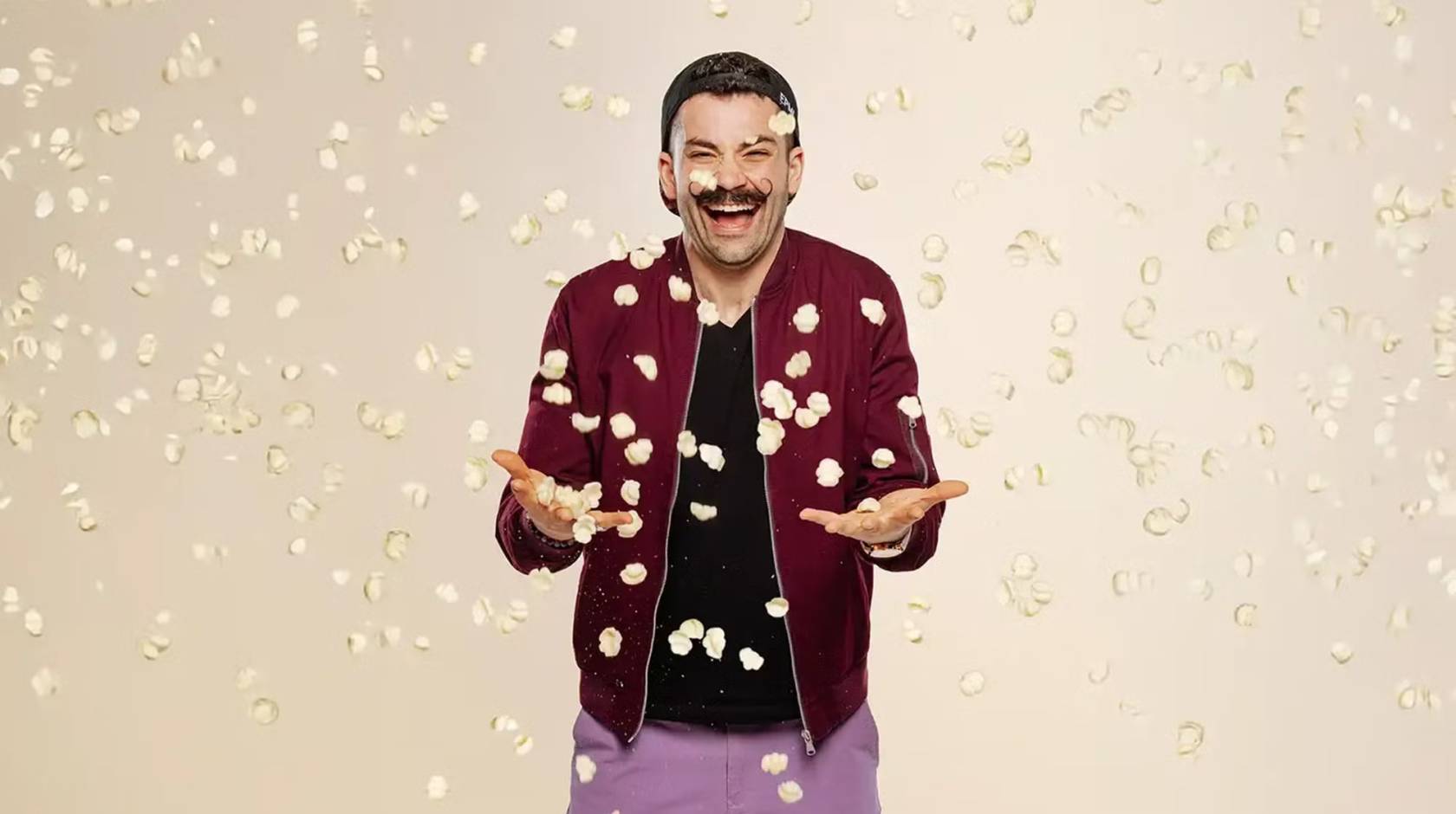 Laughing man in magneta jacket and mustache while pasta chips fall around him
