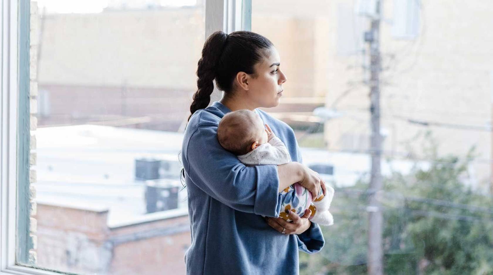 A woman with a long ponytail holds a baby and looks out the window with a trouble expression on her face