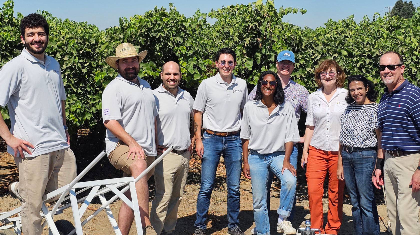 10 employees of Milano Technical Group smile for an informal photo, many wearing matching gray polo shirts, standing in a Central Valley orchard, behind a few pieces of machinery designed by the company. 