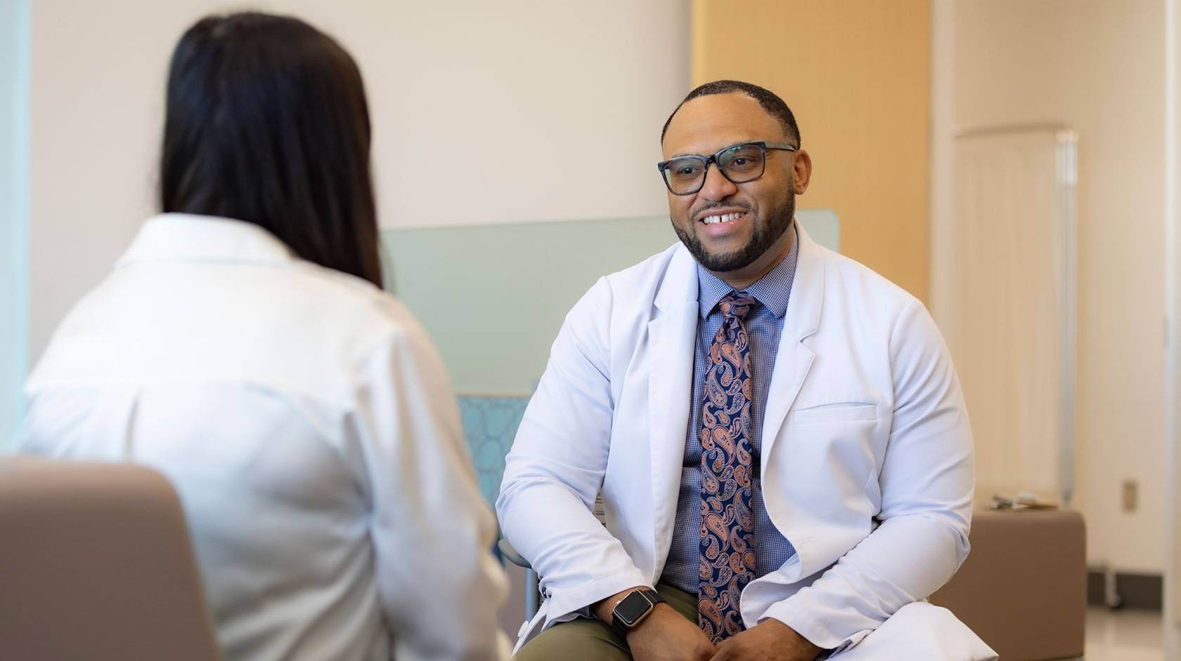 Chris Hall wearing a white medical provider's coat speaks with a patient in a lobby