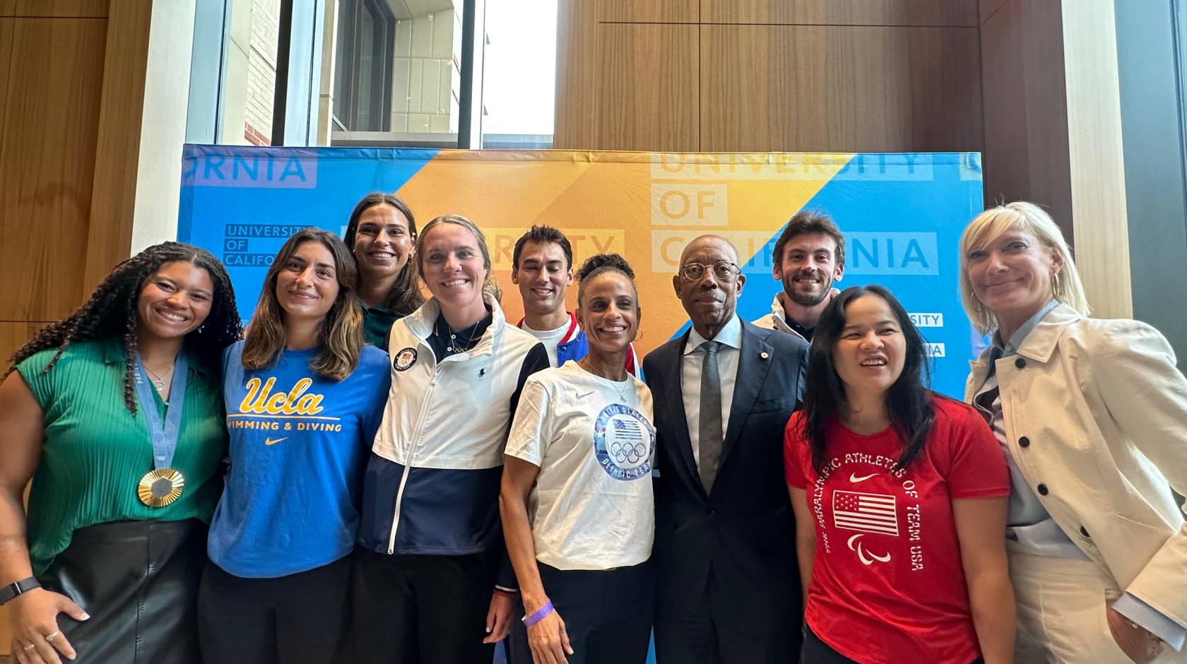 Olympian and Paralympian athletes stand with President Drake and Board of Regents Chair Janet Reilly in front of a University of California backdrop for photos
