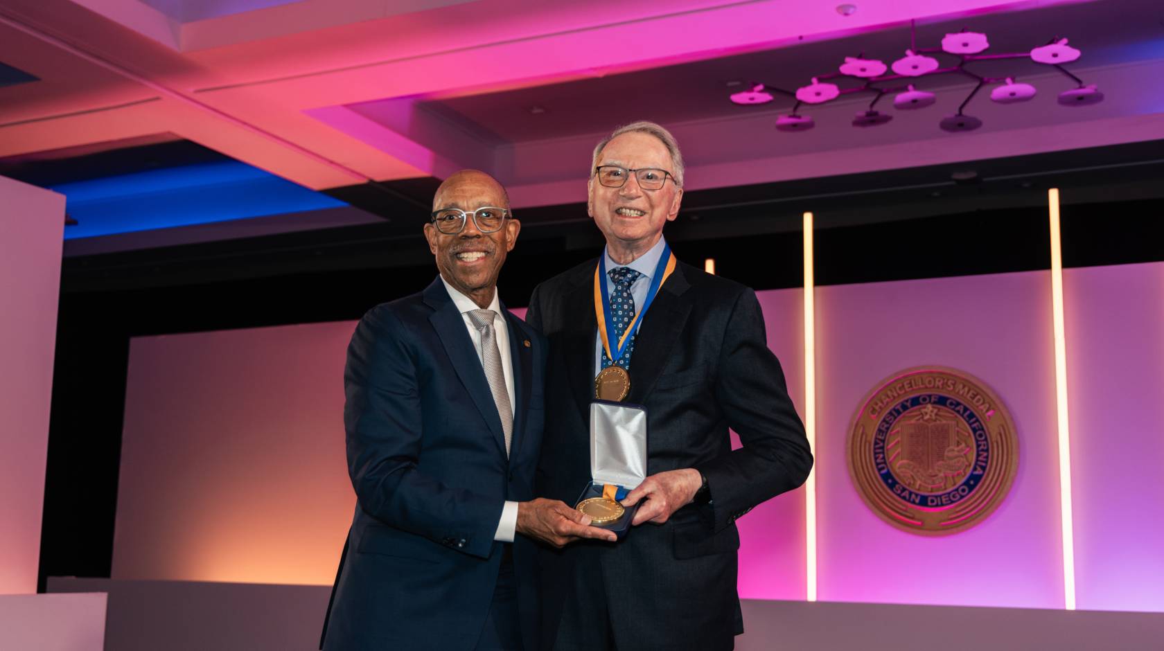 President Drake and Irwin Jacobs pose together in a room lit with purple lighting while Jacobs wears a medal and holds another