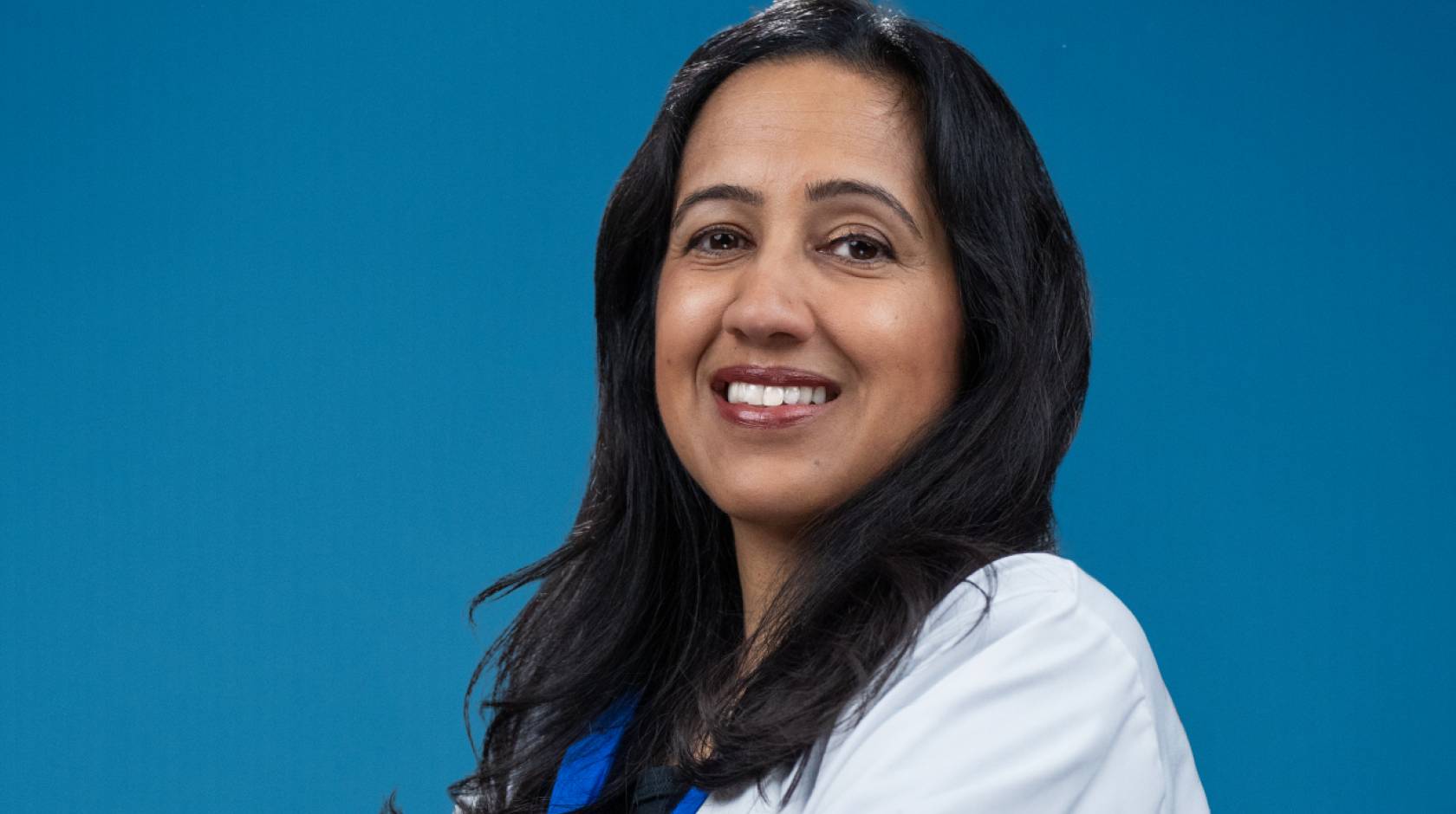 A portrait of a smiling woman with long hair in a white medical coat on a blue background