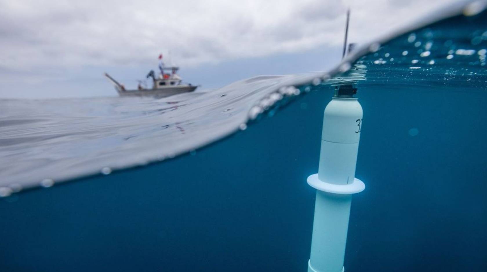 Taken with a lens that's half submerged, a photo of a cylindrical instrument floating near the surface of the ocean in the foreground, with a ship in the background
