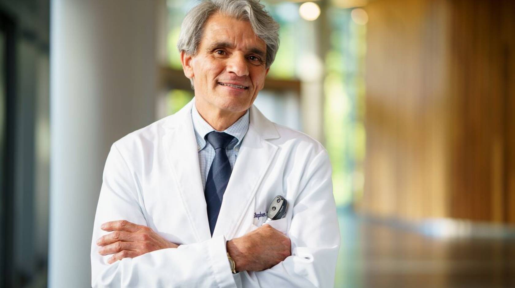 Man with white hair and arms crossed in a white doctor's coat smiles at camera in a hallway