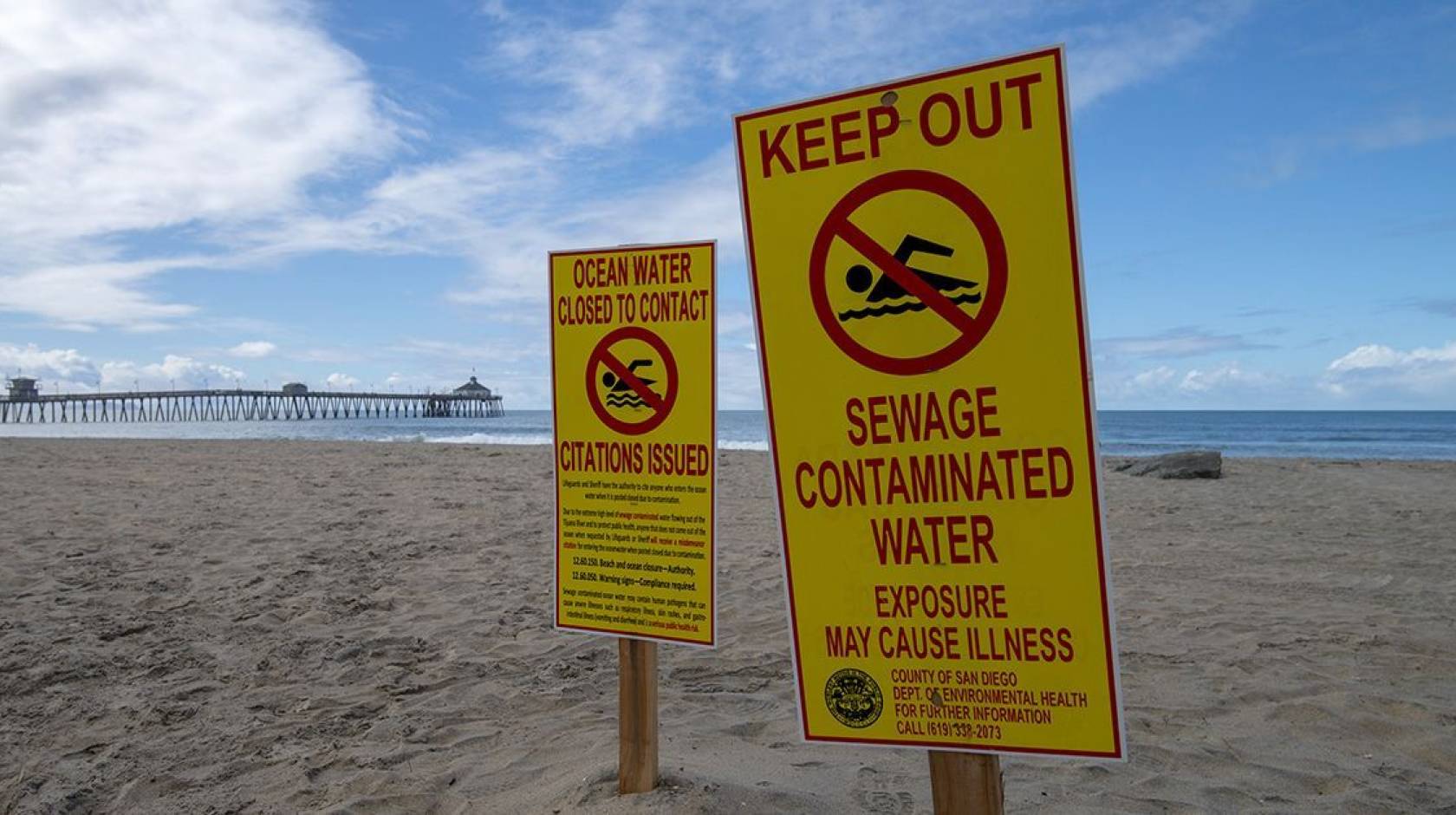 An empty beach with a pier in the background on a nice day bears signs that say Keep Out Sewage Contaminated Water and Ocean Water Closed to Contact Citations Issued