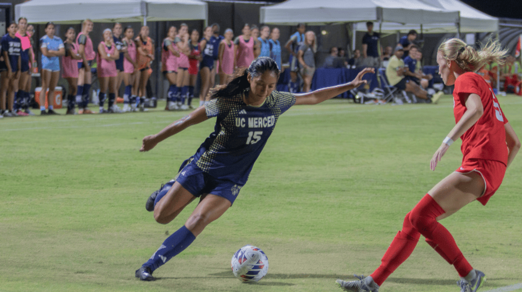 A woman in a blue UC Merced uniform prepares to kick the ball as a woman in a red uniform guards her