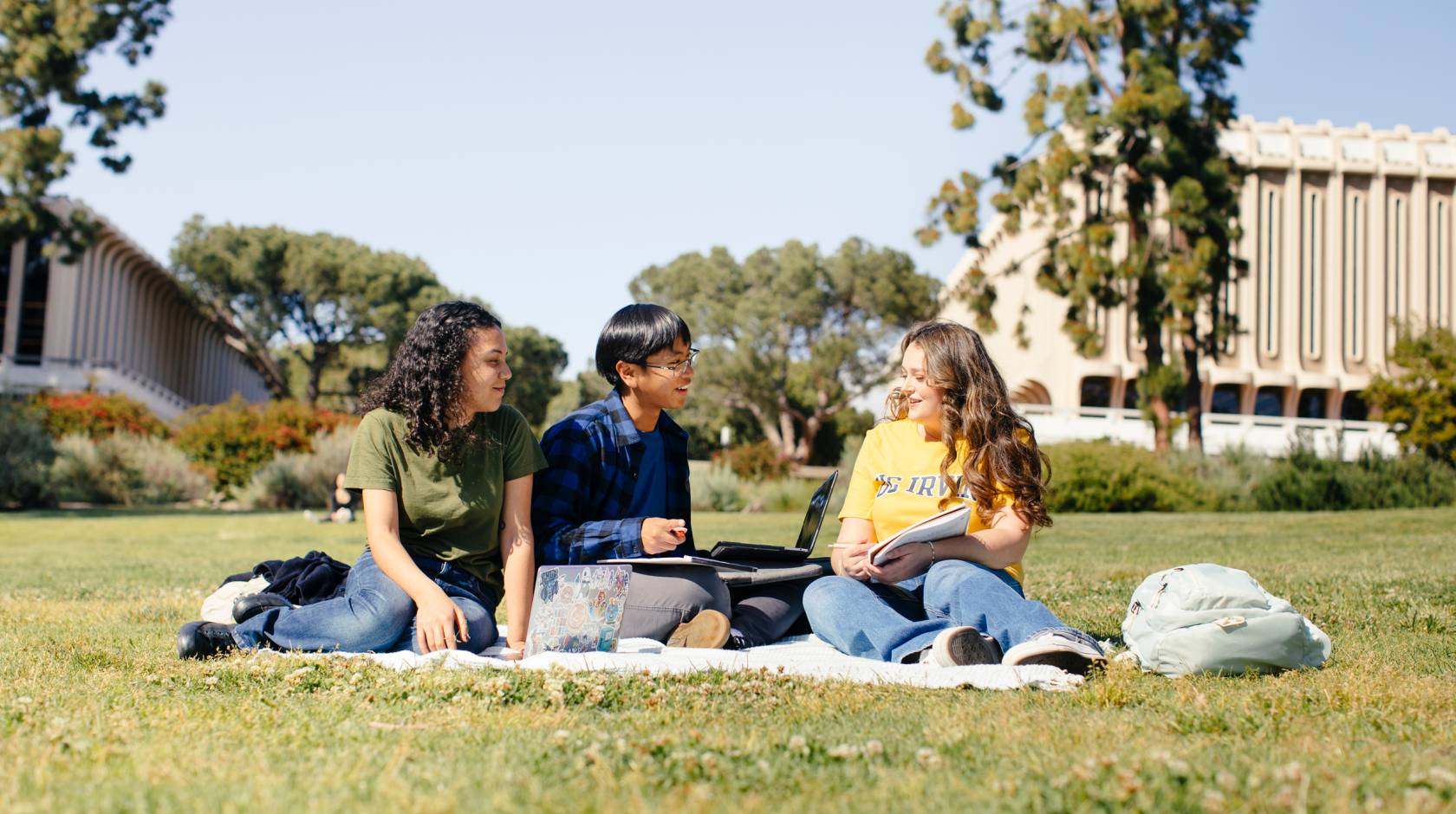 Three students on a picnic blanket studying talk to each other on the UC Irvine campus under a bright sky