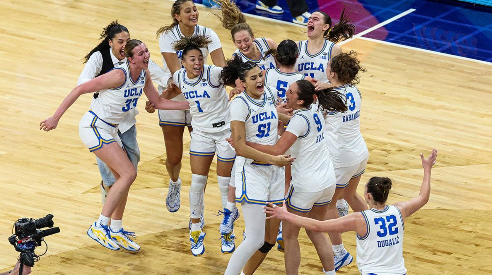 10 UCLA women's basketball players in jerseys and a woman in plain clothes jump on the court in celebration after winning the national championship
