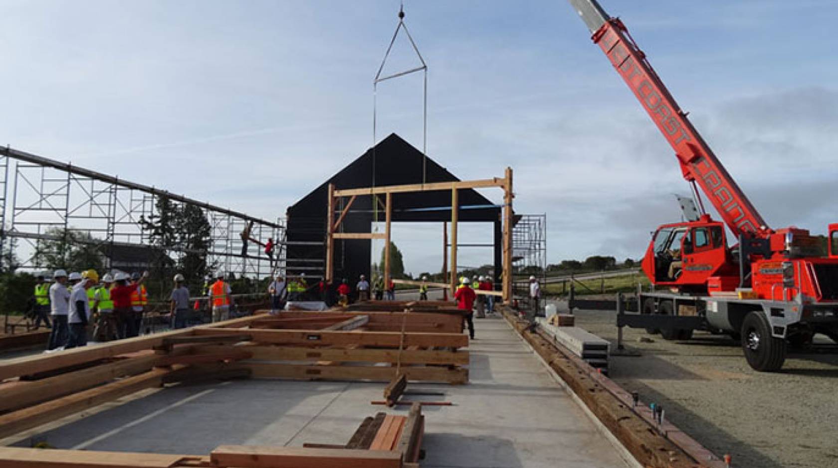 The first "bent," a cross beam, posts, and supports joined by wooden pegs, is hoisted into place by a crane early Saturday morning while others, already assembled, await their turn on the new barn's concrete slab.